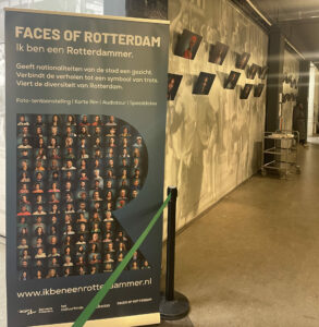 Je ziet een blauwe banner met Faces of Rotterdam als titel en daaronder een R, gevuld met allemaal kleine portretten. Daarachter zie je een muur met fotoportretten erop.