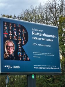 Een trotter met een blauwe achtergrond en links een grote R, vol met kleine portretjes en linksonder in de R een groter portret van een blonde dame met bril. De titel van het bord is Ik ben een Rotterdammer - Faces of Rotterdam