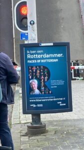 Een driehoeksbord aan een paal voor een brug met een blauwe achtergrond en in het midden een grote R, vol met kleine portretjes en linksonder in de R een groter portret van een Chinese dame met Albanisme. De titel van het bord is Ik ben een Rotterdammer - Faces of Rotterdam