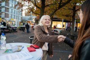 Een blonde vrouw van gemiddelde leeftijd en een bruine jas, staat bij een statafel in een winkelcentrum en geeft een jonge dame met lang, bruin haar en een bril een hand