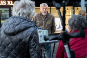 Een oudere man staat achter een statafel en praat vanuit daar met twee oudere dames met een zwarte en rode jas.