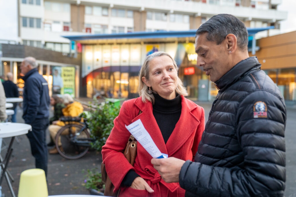 Een politicus met rood jackje praat met een burger met een bruine huidskleur, grijs haar en een zwarte jas die naar een papiertje kijkt.