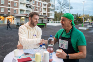 Een jongeman met een klein baardje en bruin haar in een sweater met het logo VOLT erop, leunt aan een statafel waar een fles limonade op staat, en praat met een man met bruine huidskleur en een snor en groen petje en een schort waarop staat: Aanschuiftafel