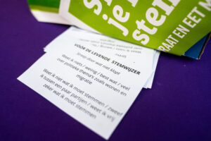 Op een paarse achtergrond ligt een flyer van de Levende Stemwijzer en een evaluatiepapiertje waarop staat 'Voor de Levende Stemwijzer', geef aan wat wel en niet klopt: weet ik weinig'best wat ' veel over politieke thema's zoals wonen en migratie. En dan een witregel en dan staat er: Wist ik niet wat ik moet stemmen / zweef ik tussen een paar partijen / weet ik vrij zeker wat ik moet stemmen.