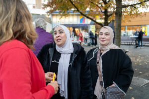 Twee vrouwen met een witte en beige losse hoofddoek en zwarte jassen praten met een vrouw met een rood jasje.