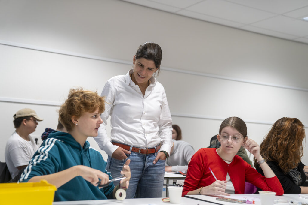 Je ziet een jongere links met oranje haar en een blauw vest en rechts met bruin haar en een rood vestje en in het midden een volwassene in spijkerbroek met witte blouse die luistert.