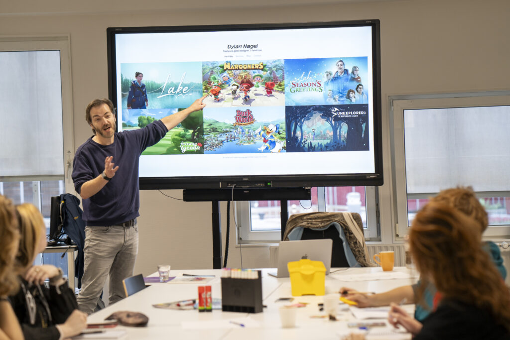 Een witte man met spijkerbroek en blauwe trui wijst naar een scherm waar plaatjes van games op te zien zijn