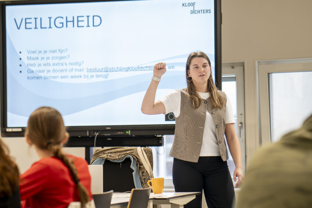 Een witte vrouw met wit shirt en lange bruine harden doet haar hand omhoog met vuist in een klas. Achter haar zie je een powerpointscherm met de titel 'Veiligheid'.