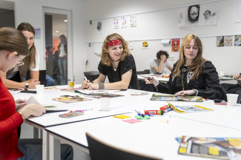 Aan een witte tafel vol met post-its en pritstiften en tijdschriften in een klas zitten een jongen met lang blond haar en een rode bandana met een zwart shirt en rechts van hem een meisje met half blond en half bruin haar met een zwart vest.