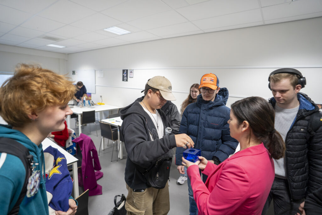 Een groep jongeren staat om een vrouw met een roze blazer en donkerbruin haar heen. Een jongere stopt iets in een blauw doosje.